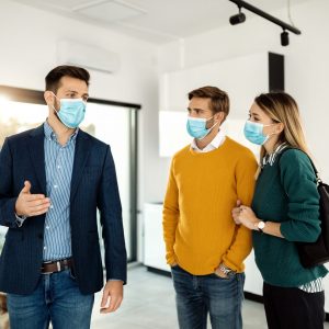 Real estate agent talking to a couple while showing them a new apartment during coronavirus pandemic. Young couple and male agent wearing face masks while looking a new real estate.